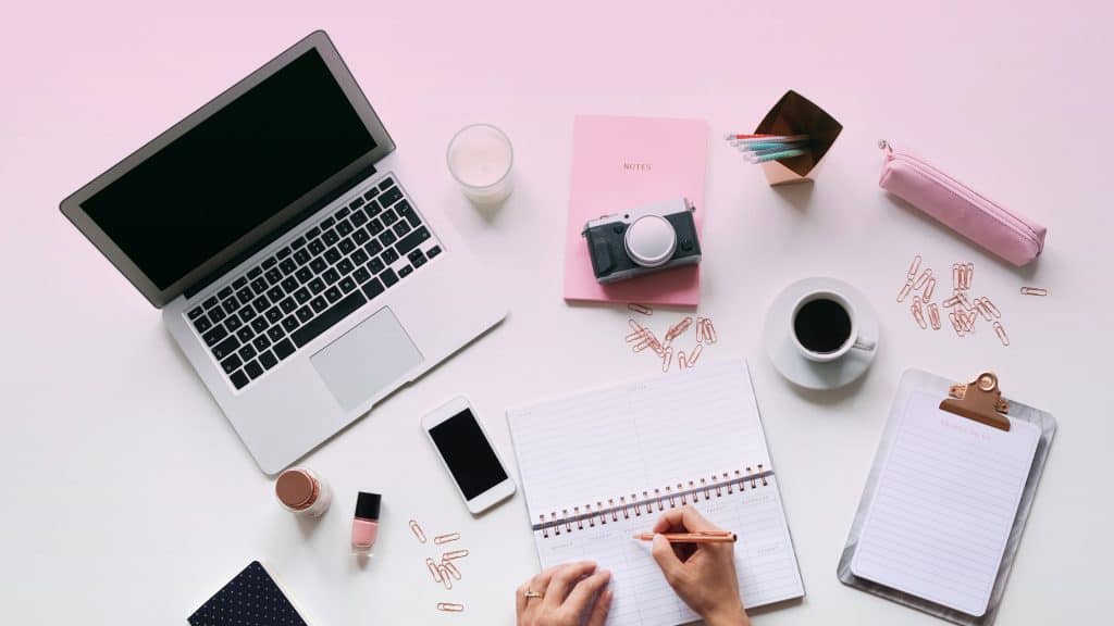 A feminine flat lay workspace featuring a silver laptop, pink notebook with a camera on top, a phone, open planner, pens, coffee, candles, and rose gold paper clips on a white desk with a soft pink gradient background. A hand is seen writing in the planner.