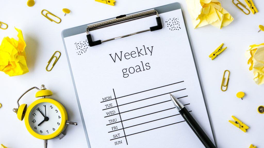 A clipboard with a weekly goals sheet labeled Monday through Sunday, surrounded by a bright yellow alarm clock, yellow paper clips, thumbtacks, crumpled notes, and clothespins. A pen rests on the paper, suggesting planning in progress.