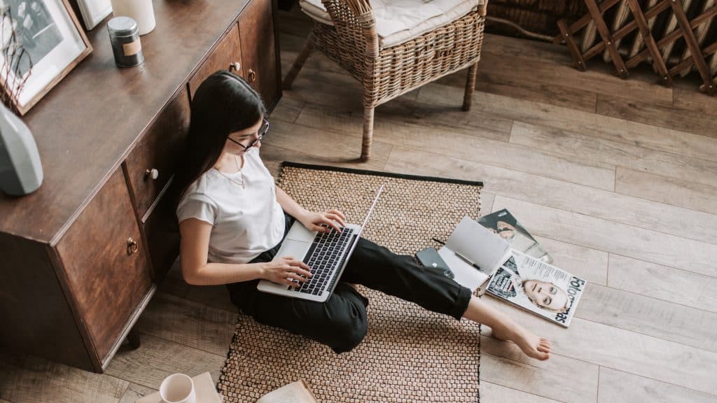 A woman with glasses sits on the floor using a laptop, leaning against a wooden cabinet. Around her are open notebooks, magazines, a phone, and a coffee mug on a cozy woven rug in a homey setting with wicker and wood furniture.